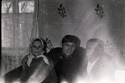 A candid portrait of three people inside a home, likely Bulgaria, 1960s-1970s. An older woman in a headscarf sits flanked by ...