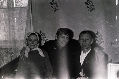 Three generations pose in a modestly decorated interior. A woman in a headscarf, a young man in a suit jacket, and an older m...