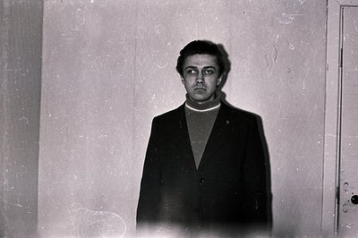 A man stands centered against a textured wall, wearing a dark suit and turtleneck sweater. The image, likely a portrait, exhi...