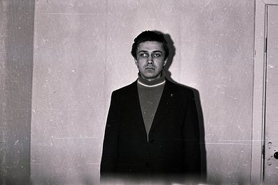 Striking black and white portrait of a man in a dark suit and turtleneck sweater, set against a textured wall. Direct gaze, m...
