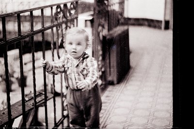 A young boy, likely a toddler, stands near a wrought iron gate in a courtyard setting. He wears a plaid shirt and overalls. T...
