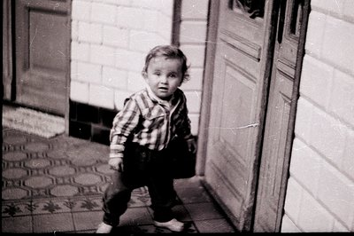 A young child in short overalls & checked shirt stands poised on a tiled entryway. Textured wall & weathered door suggest a r...