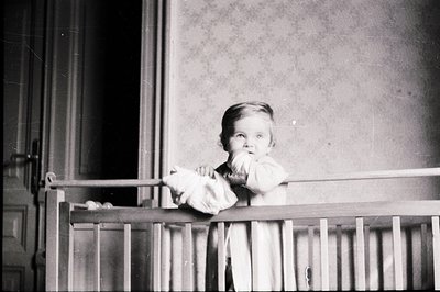 Young child peering over a crib railing. Interior scene features patterned wallpaper and a partially visible doorway. Likely ...