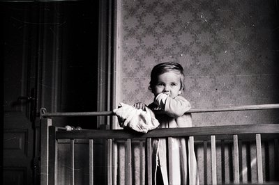 A young child stands at a balcony railing, clutching a plush toy. The image displays a weathered architectural detail with in...