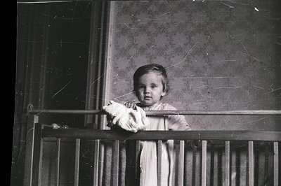 A young child, possibly a toddler, stands peering from behind a wooden balcony railing. The child wears a simple, light-color...