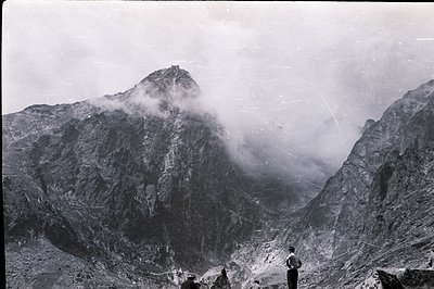Dramatic alpine landscape with a towering peak partially obscured by swirling clouds and mist. Three figures are positioned i...