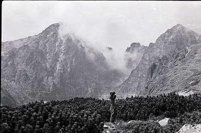 Monk silhouetted against a dramatic, mist-shrouded mountain range. Dense coniferous forest fills the foreground, framing the ...