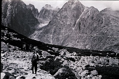 A lone figure stands amidst a rocky alpine landscape, facing towering, jagged mountains. The scene appears sparsely vegetated...