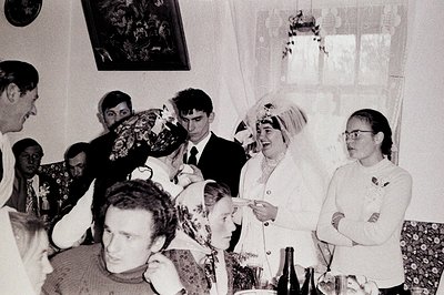 Black & white photo captures a wedding reception scene. A bride in a white suit & headscarf stands with her groom, surrounded...