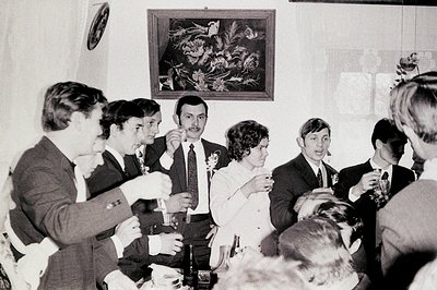 A candid shot at an indoor gathering, likely a wedding reception. Six individuals are visible, dressed formally in 1960s atti...