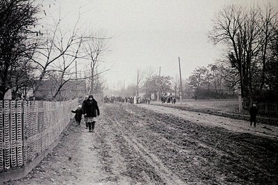 A group of people walks along a muddy, unpaved road, likely refugees or displaced persons. A tall, ornamental wooden fence li...