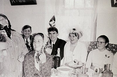 A group of five people, dressed in formal attire and festive headwear, are seated around a table laden with food. The scene a...