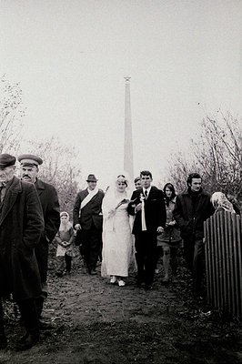 A bride and groom, formally dressed, walk alongside their wedding party with a tall Soviet memorial (likely a monument to WWI...
