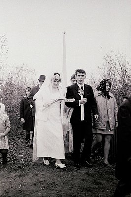 A formally dressed wedding couple stands center frame, posed outdoors, facing a stark, imposing Soviet-era monument featuring...