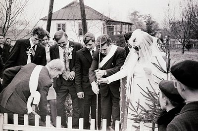 A wedding ceremony outdoors, possibly a tree planting ritual. A formally dressed couple, veiled bride and suited groom, are a...
