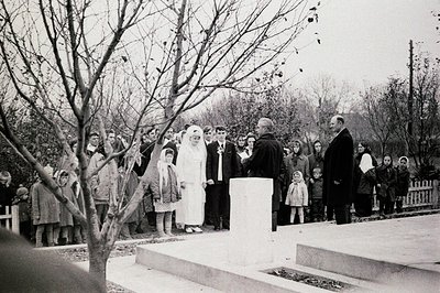 A solemn ceremony outdoors. A bride and groom, center, are flanked by a group of formally dressed attendees, including childr...