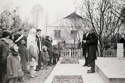 A formal gathering, likely a memorial service, takes place outside a modest, single-story building. A man in a dark coat and ...