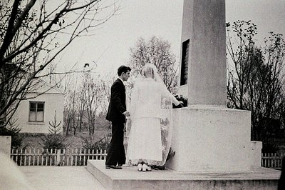 A formally-dressed couple stands before a monumental stone structure, likely a memorial. The bride wears a long-sleeved, high...
