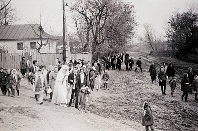 A wedding procession walks along a dirt road, likely in a rural area. The bride wears a traditional veil, accompanied by a gr...