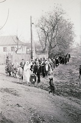A wedding procession moves along a rural, unpaved road. The bride, in a traditional gown & veil, is surrounded by formally dr...