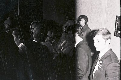 A candid black and white photo captures a moment during what appears to be a formal event. Three men in suits, and a woman wi...