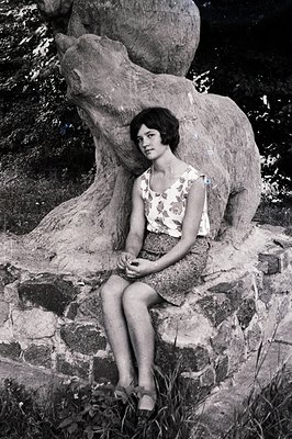 A young woman sits posed near a textured, large tree root sculpture forming a stylized bench. She wears a floral-print top an...