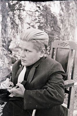 Formal portrait of a man seated in a decorative chair, looking off-camera. He wears a dark suit, tie, and has a distinctive, ...