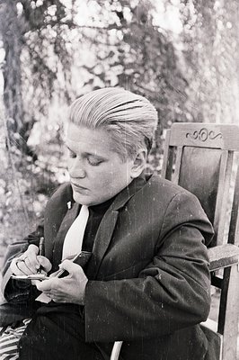A man in a dark suit sits writing in a small notebook, outdoors. He has a distinctive, slicked-back hairstyle and is wearing ...