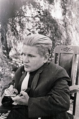 A formally dressed young person, likely a teenager, sits outdoors writing with a fountain pen. The subject wears a dark suit ...