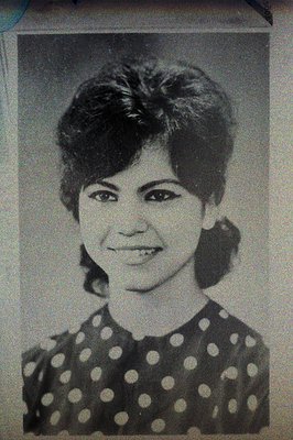 Black and white studio portrait of a young woman with heavily styled hair and distinct eye makeup. She wears a dark top with ...