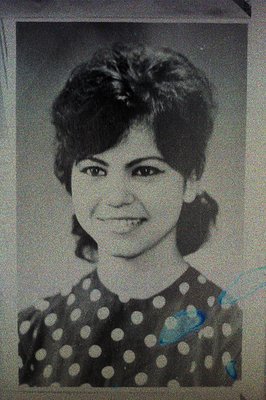 A black and white portrait shows a young woman with a short, layered hairstyle and dark eye makeup. She wears a polka-dot top...