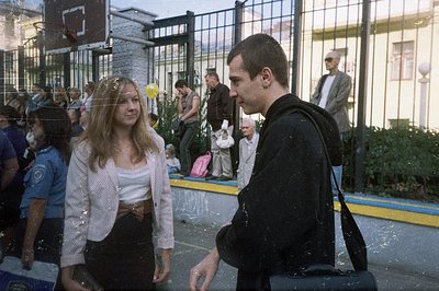 A young couple stands facing each other in a courtyard setting, possibly a public event or festival. A uniformed officer stan...