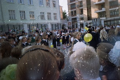 A group of children in formal wear and white hats gather outdoors, likely a school event or graduation. Buildings with distin...