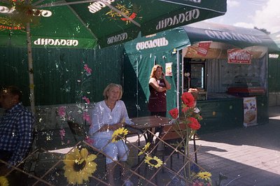An outdoor scene shows a woman seated at a cafe table adorned with sunflowers. A second woman stands behind a food kiosk, dis...