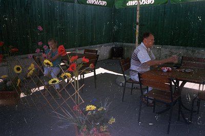 Outdoor scene depicting a young girl and an older man seated at tables in a shaded courtyard. Vibrant sunflowers & floral arr...