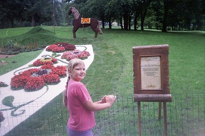 A young girl in a pink t-shirt stands beside a wire fence, facing the camera. Behind her, a tiered floral display features re...