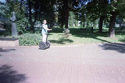 A person rides a Segway scooter on a brick-paved path within a park. A statue and a low brick wall are visible on the left. L...