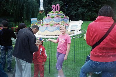 A young girl in a pink shirt and purple leggings stands before a large floral sculpture resembling a tiered cake with "2011" ...