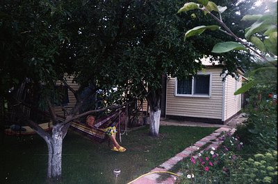 A woman relaxes in a hammock strung between trees in a lush, green garden. A modest, light-colored building with a single win...