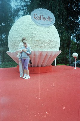 A young girl and woman stand before a large, sculpted confection resembling a Raffaello candy, branded with the name "Confeit...