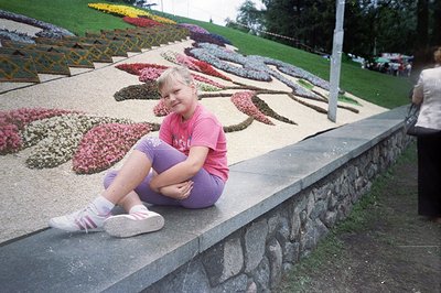A young girl sits on a stone wall, viewing a meticulously arranged flowerbed. The floral display features concentric pink and...