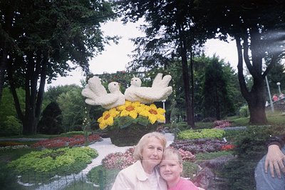 A color photograph depicts a woman and young boy positioned in front of elaborate floral topiaries. The figures are framed by...