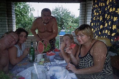 A family gathering under a covered patio, possibly in Bulgaria, features a man shirtless & cutting a watermelon. Three women ...