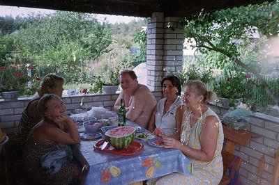 A family gathers around a table covered with a floral tablecloth, enjoying a watermelon feast. The setting is a vine-covered ...