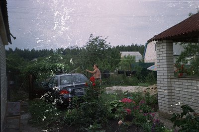A man washes a dark blue sedan in a leafy, residential garden. Brick and stone structure with red roof visible. Laundry hangs...