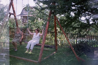 Two women enjoy a swing set in a leafy, fenced-in yard. The scene evokes a casual, domestic moment, likely captured in the 19...
