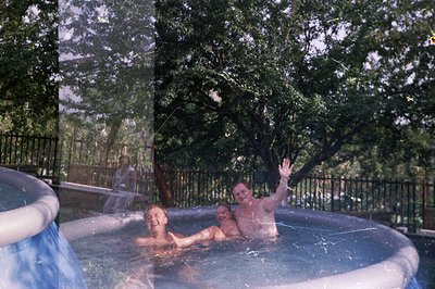 Two men relax in an inflatable hot tub. One raises a hand in greeting. A wooden deck, fence, and lush foliage form the backdr...