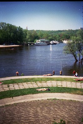A riverside scene; people relax on the grass and pavement alongside a wide river, potentially the Danube. Boats are visible a...