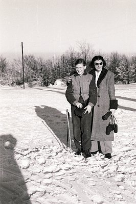 Two figures in winter outerwear stand in a snowy landscape. The woman, in a long coat & dark sunglasses, carries a handbag. T...