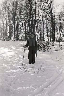 A young person stands in a snow-covered landscape, holding a walking stick. The figure is bundled in outerwear, appearing to ...
